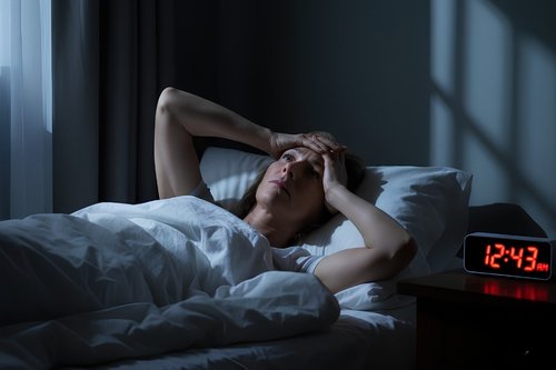 A woman lying in bed at night looking stressed and awake, with a digital alarm clock showing 12:43 AM, illustrating common sleep problems after 40.