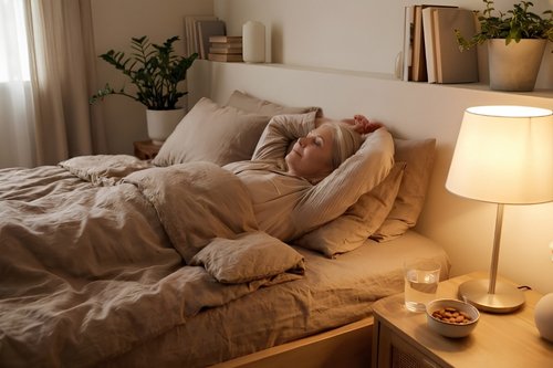 A middle-aged woman with grey hair is sleeping soundly in a bed with linen sheets, her arms tucked behind her head. A glowing lamp is on the bedside table next to a glass of water and a bowl of almonds.