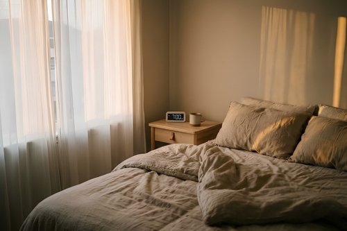 A cozy, sunlit bedroom with a made bed and a digital clock showing 7:00 AM, illustrating healthy sleep hours after 25.